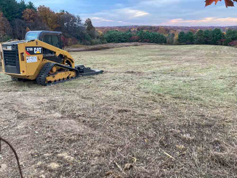 A scene of land clearing or cleanup in a wooded area. A bonfire is burning in the center, sending up plumes of bluish-gray smoke, possibly from the burning of green or moist plant material. A yellow mini excavator is positioned to the right, likely used for moving debris or assisting in the clearing process.