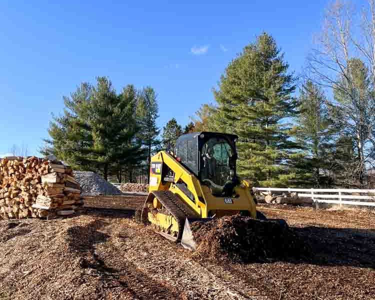 A compact yellow skid steer loader in a rural setting with a clear blue sky above. The machine is sitting on a bed of mulch, which could suggest recent landscaping or land-clearing work.