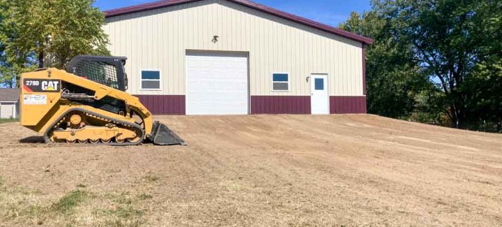 A yellow compact track loader is parked on a dirt terrain that appears to be recently graded in preparation for landscaping or construction. The sky is clear and blue, suggesting a sunny day. The area is devoid of grass, possibly due to recent construction or landscaping activity.