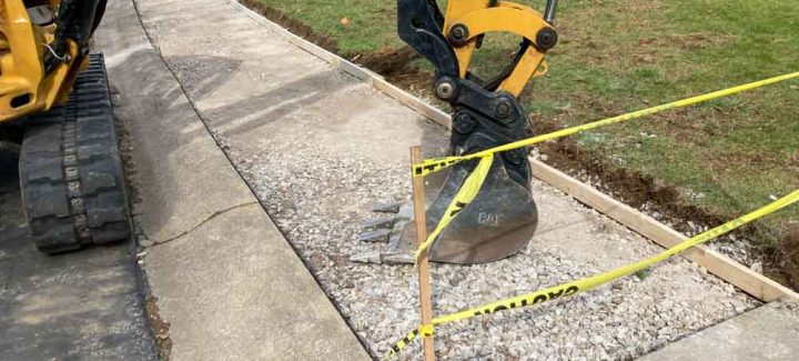 This image features a close-up view of a construction or repair site, with a focus on a section of sidewalk where work is being done. Part of a yellow excavator arm is visible on the left, with its bucket positioned close to the ground, ready for operation. The excavator is possibly being used to dig or adjust the gravel foundation in preparation for laying concrete. There's a wooden form set up to define the boundaries of the new section of sidewalk that needs to be poured. Caution tape is strung around the area to mark the work zone and ensure safety, warning passersby to keep a distance. The existing sidewalk next to the work area looks slightly worn, suggesting this may be a repair or improvement project.