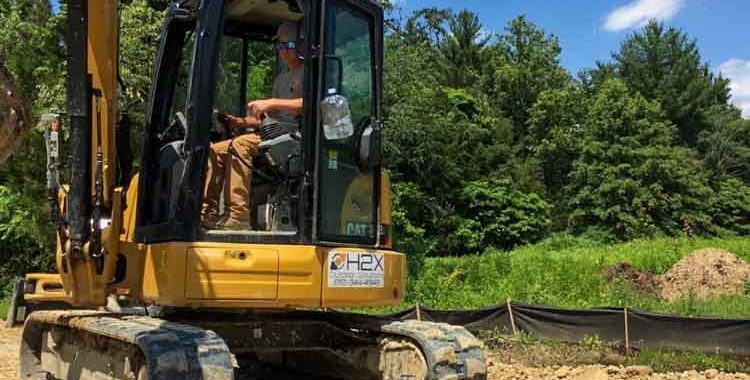 A close-up of a yellow hydraulic excavator on a construction site with a clear, blue sky overhead. The operator is inside the cabin, which suggests that the machine is either in operation or about to be used. The excavator is positioned on a bed of rough, uneven dirt, and there are foundation structures in the form of wooden forms and rebar, indicating that concrete will be poured for foundations or slabs. In the background, a pile of earth and black silt fencing can be seen, typical of construction sites to control sediment run-off.