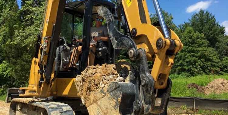 An excavator lifting a mass of soil with rocks, digging out a trench for possible drainage system.