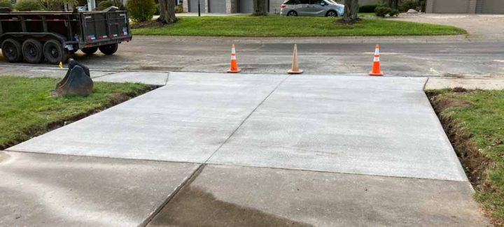 A new, smooth concrete driveway with expansion joints, recently installed in a residential neighborhood. Houses with driveways and yards are visible in the background.