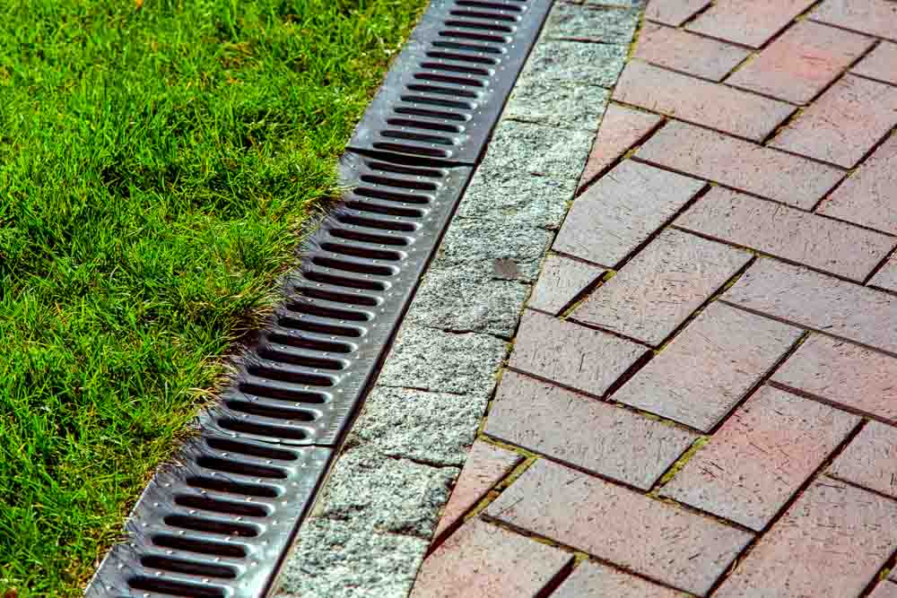 An intricate iron drainage system seamlessly integrated between sleek paving slabs and lush green lawn, ensuring efficient water flow and a harmonious blend of urban design and natural elements.